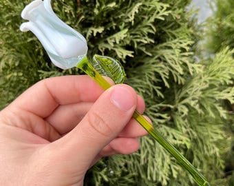 May include: A handcrafted glass flower sculpture featuring a white and light blue tulip bloom. The stem and leaf are a vibrant green. The glass art piece is held against a backdrop of green foliage.