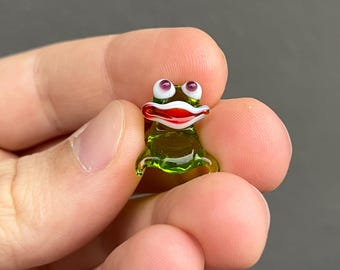 May include: A small, green glass frog ornament with red lips, white detailing, and purple eyes. The frog is held between fingers against a grey background. The frog's body is translucent, and its features are detailed.