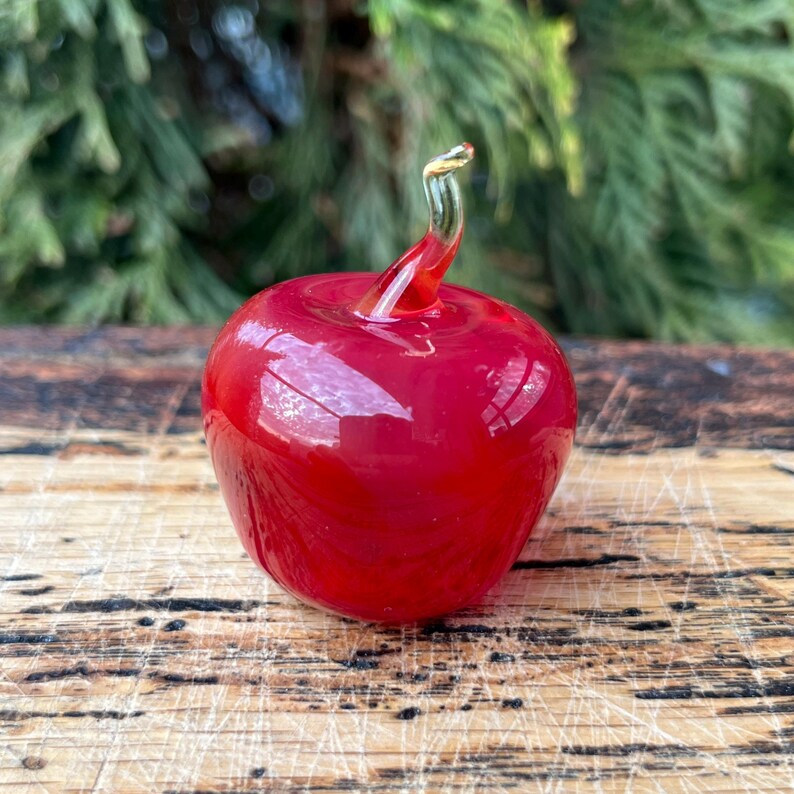 May include: A small, smooth, bright red glass apple figurine with a clear, curved glass stem extending from the top. The apple has a glossy, reflective surface that catches the light, showcasing a vibrant, solid red color throughout the body.