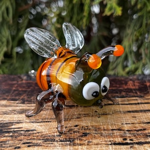 Puede incluir: Una figura de abeja de vidrio hecha a mano con alas transparentes, antenas naranjas y abdomen a rayas. El cuerpo de la abeja presenta una combinaci&oacute;n de tonos verdes, naranjas y amarillos. La figura est&aacute; sobre un fondo verde borroso.