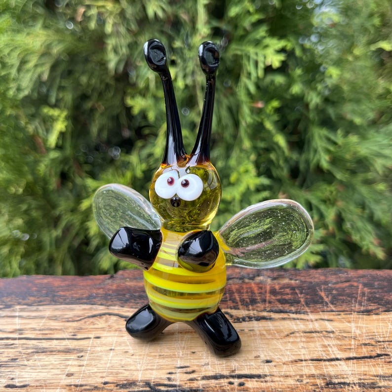 Puede incluir: Figura de abeja de cristal hecha a mano con cuerpo a rayas amarillas y negras, alas transparentes y antenas negras. La abeja tiene grandes ojos blancos con pupilas rojas. La figura se exhibe sobre una superficie de madera.