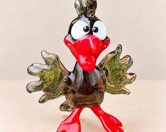 May include: A glass figurine of a bird with a dark brown body, translucent olive green wings, and bright red feet and beak. The bird has large white eyes with black pupils, a crest on its head, and is standing in an upright, alert position.