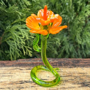 Puede incluir: Una escultura de flor de vidrio con una flor naranja y un tallo verde. El tallo está enroscado en un bucle en la base.