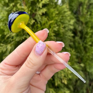 May include: A glass mushroom sculpture with a yellow cap and a blue base. The stem is clear glass and is long and thin.
