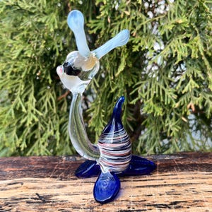 May include: A handcrafted glass sculpture of a seal. The seal has a blue base and flippers, a striped body with red, white, and black accents, and a clear head with light blue appendages. The sculpture is set against a backdrop of green foliage.