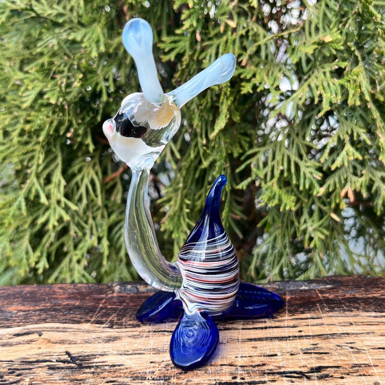 May include: A handcrafted glass sculpture of a seal. The seal has a blue base and flippers, a striped body with red, white, and black accents, and a clear head with light blue appendages. The sculpture is set against a backdrop of green foliage.