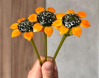 Puede incluir: Tres esculturas de girasoles de vidrio con pétalos de color naranja vibrante y centros oscuros moteados. Cada flor está unida a un tallo de vidrio verde translúcido y delgado, creando un conjunto coherente de piezas de arte botánico.