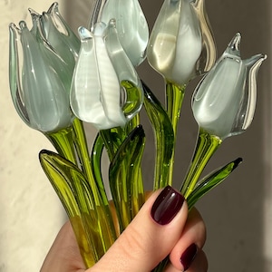 May include: A bouquet of glass tulips in shades of white and pale blue, with green stems and leaves. The flowers are held in a hand with burgundy nail polish. The glass is translucent, allowing light to pass through.