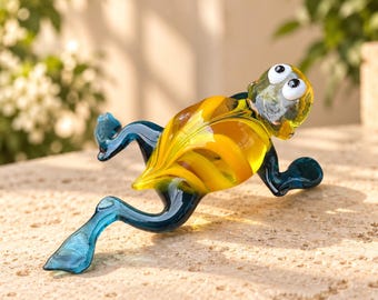 Könnte beinhalten: Eine Glasfrosch-Figur mit einem durchscheinenden gelb-orangen Körper und dunkelblauen Gliedmaßen. Der Frosch hat zwei hervorstehende weiße Augen mit schwarzen Pupillen und einen Körper aus glattem, geschwungenem Glas in liegender Pose.
