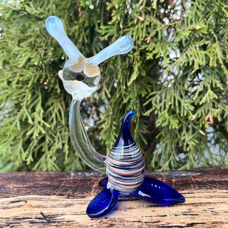 May include: A whimsical glass sculpture of a seal. The seal features a clear body with a striped torso in shades of white, black, red, and blue. The flippers and head are a vibrant blue, and the head has two light blue antennae.