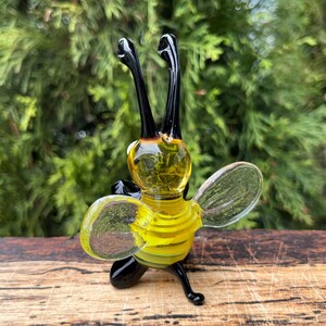 Puede incluir: Una figura de abeja de vidrio hecha a mano con rayas amarillas y negras en el cuerpo, alas transparentes y antenas y patas negras. La abeja est&aacute; colocada en posici&oacute;n vertical sobre una superficie de madera, con un fondo verde borroso.