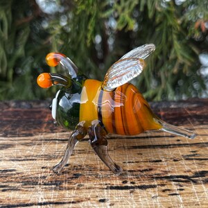 Puede incluir: Figura de abeja de cristal hecha a mano con rayas naranjas, amarillas y marrones. La abeja tiene alas transparentes, antenas naranjas y patas oscuras. La figura se exhibe sobre una superficie de madera, mostrando los intrincados detalles.