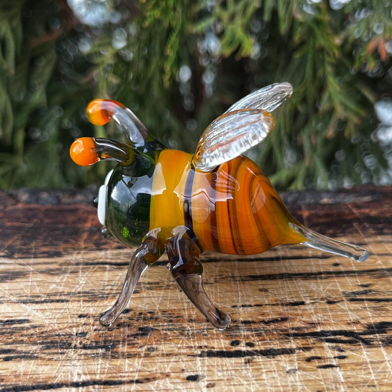 Puede incluir: Figura de abeja de cristal hecha a mano con rayas naranjas, amarillas y marrones. La abeja tiene alas transparentes, antenas naranjas y patas oscuras. La figura se exhibe sobre una superficie de madera, mostrando los intrincados detalles.