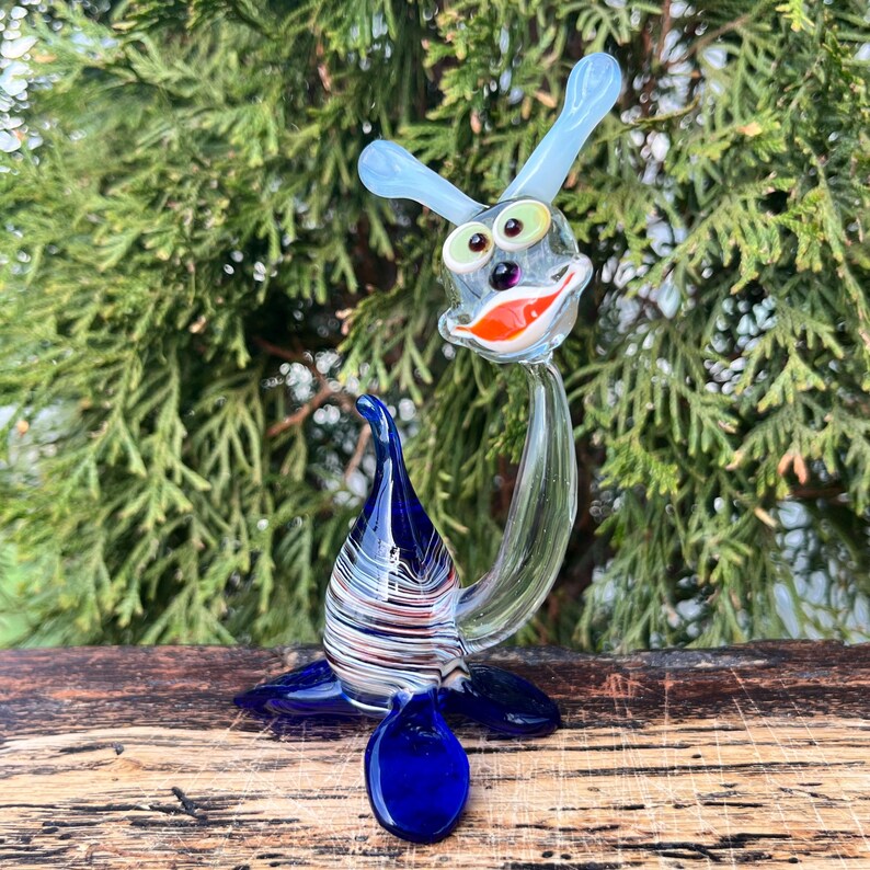 May include: A whimsical glass sculpture of a seal-like creature. The sculpture features a blue body with white and red stripes, a clear neck, and a head with blue antennae, green eyes, and an orange and red mouth. The base is dark blue.