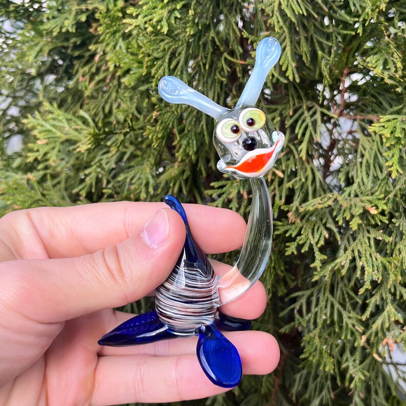 May include: A whimsical glass sculpture of a sea creature with a smiling face, blue fins, and light blue antennae. The body is a mix of clear, blue, and white glass. The creature's face has green eyes and a red smile.