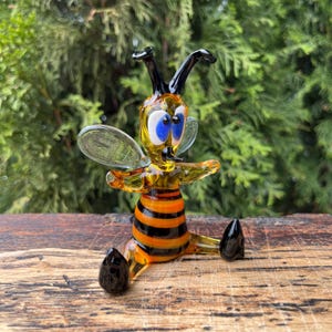 May include: A whimsical glass bee figurine with a yellow body, black and orange striped abdomen, and large blue eyes. The bee has clear wings and black antennae and feet. It is posed sitting on a wooden surface.