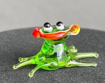 May include: A small green glass frog figurine with red and orange accents on its mouth. The frog has black eyes and white pupils. The figurine is sitting on a black surface.
