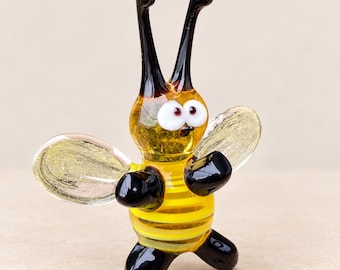 Puede incluir: Una figura de cristal de una abeja con cuerpo a rayas amarillas y negras, dos largas antenas negras y dos ojos blancos con pupilas oscuras. La abeja tiene alas transparentes texturizadas y pies de cristal negro, en posición erguida.