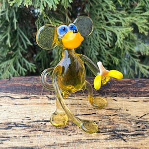 May include: A handcrafted glass monkey figurine in shades of yellow and clear. The monkey has large ears, blue eyes, and a yellow mouth. It holds a yellow and white banana. The figurine is posed on a wooden surface.