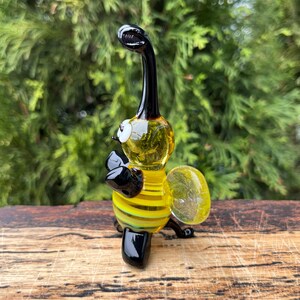Puede incluir: Una figura de abeja de vidrio hecha a mano. La abeja presenta un cuerpo a rayas amarillas y negras, un ala amarilla transparente y una cabeza y patas negras. La figura se encuentra sobre un fondo verde borroso.