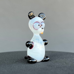 Puede incluir: Una pequeña figurita de vidrio de un oso panda. El panda es blanco con orejas y patas negras. Tiene ojos grandes y redondos y una pequeña nariz negra.