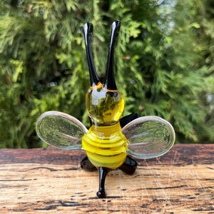 Puede incluir: Figura de abeja de cristal hecha a mano con rayas amarillas y negras en el cuerpo y alas transparentes. La abeja tiene dos antenas negras y est&aacute; sobre una superficie de madera.