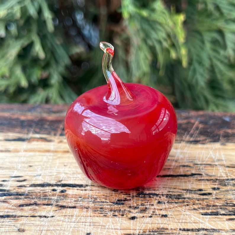 May include: A small, solid glass apple figurine in a vibrant, translucent red hue. The object features a smooth, rounded body and a clear, curved glass stem extending from the top, showcasing a glossy finish and reflective surface quality.