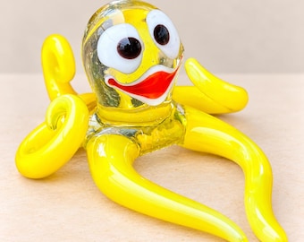 Può includere: Una statuetta di polpo in vetro con un corpo giallo brillante e una finitura liscia e lucida. La creatura presenta due grandi occhi bianchi con pupille scure e una bocca rossa prominente. Diversi tentacoli gialli ricurvi si estendono.