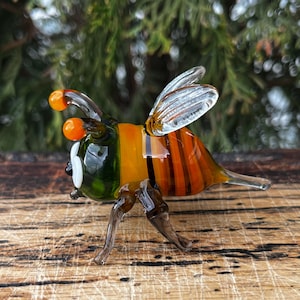 Puede incluir: Una figura de abeja de cristal hecha a mano. La abeja presenta un cuerpo a rayas naranja y marr&oacute;n, alas transparentes y antenas naranjas. La figura se encuentra sobre un fondo verde borroso, lo que sugiere un entorno al aire libre. Las patas de la abeja tambi&eacute;n son de cristal transparente.