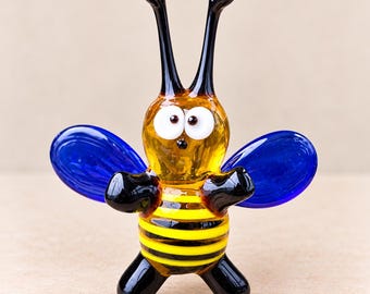 May include: A glass figurine of a bee with a yellow and black striped body, two large white eyes with black pupils, long black antennae, and two translucent blue wings. The bee has black arms and legs and a clear amber-colored head section.