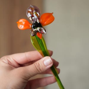 Op de afbeelding: Een decoratief glazen bloemornament met een doorschijnend paars bolvormig midden gevuld met veelkleurige spikkels, twee levendige oranje bloemblaadjes en een lange, gladde, felgroene steel die onderaan in een punt uitloopt.