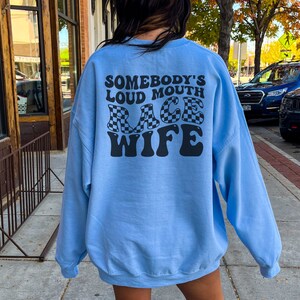 May include: Light blue sweatshirt with the text "Somebody's Loud Mouth Race Wife" in black lettering with a checkered flag design.