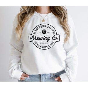Puede incluir: Sudadera blanca con un logotipo en blanco y negro que dice "Sanderson Sisters Brewing Co." con un caldero y el texto "Est. 1693" y "Salem, Mass. Premium Witches Brew."