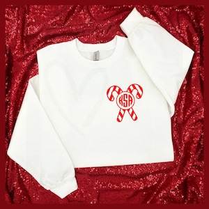 May include: White sweatshirt with a red candy cane design. The candy canes have red and white stripes and a monogram in the center. The sweatshirt has long sleeves and a crew neck.