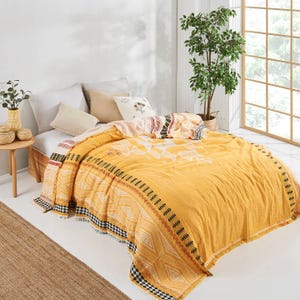 May include: A yellow and white patterned quilt with fringe on the edges. The quilt is on a bed with white pillows and a brown wooden nightstand.