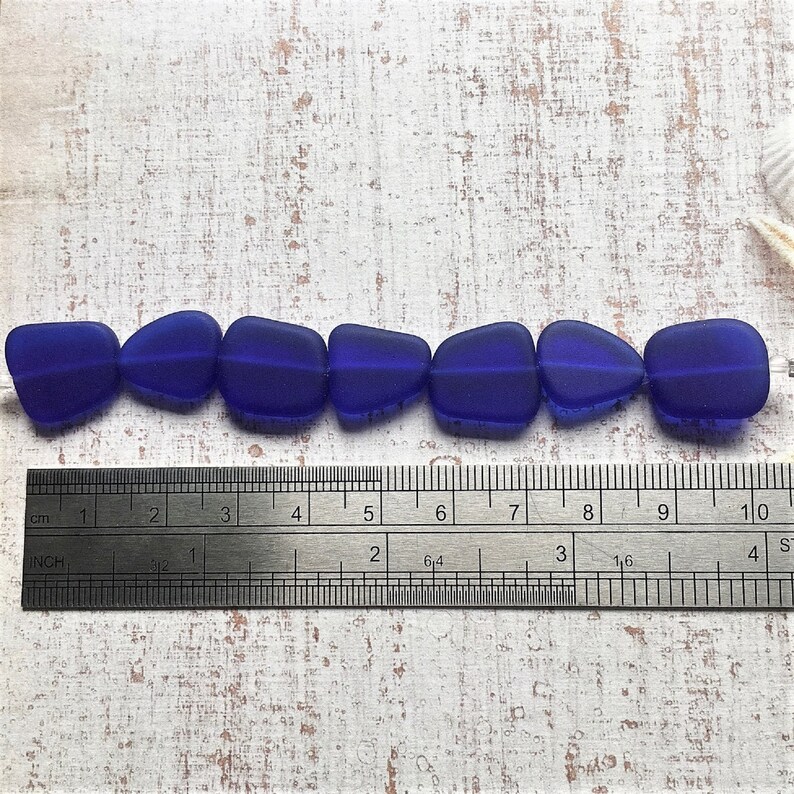 K&ouml;nnte beinhalten: Eine Reihe von sieben blauen Glasperlen, jede mit einer leicht unterschiedlichen Form, auf einer wei&szlig;en Oberfl&auml;che neben einem Lineal in Zentimetern und Zoll.