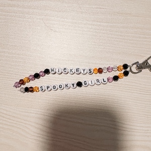 May include: A beaded keychain featuring two strands of white letter beads spelling MICKEYS and SPOOKY GIRL, interspersed with faceted black, orange, and purple beads. The strands are connected to a silver-toned metal lobster claw clasp.