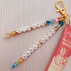 May include: A gold-toned keychain featuring two beaded strands with white letter beads spelling SPF AND PIXIE DUST, accented by colorful translucent beads, attached to a tube of Pink Pineapple Sunrise lip gloss labeled .47 FL OZ.