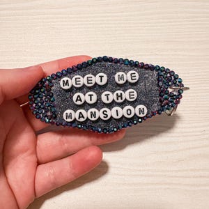 May include: An oval-shaped keychain featuring a dark gray glittery surface bordered by small, iridescent dark blue and purple rhinestones. The text "MEET ME AT THE MANSION" is spelled out in white plastic beads with black lettering across the center.