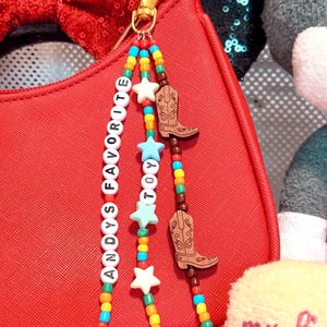 K&ouml;nnte beinhalten: Ein Taschenanh&auml;nger mit drei Perlenstr&auml;ngen an einem goldfarbenen Karabinerverschluss. Die Str&auml;nge bestehen aus bunten Kunststoffperlen, wei&szlig;en Buchstabenperlen mit der Aufschrift ANDYS FAVORITE TOY, hellblauen und wei&szlig;en sternf&ouml;rmigen Perlen sowie zwei braunen Holzanh&auml;ngern in Form von Cowboystiefeln.