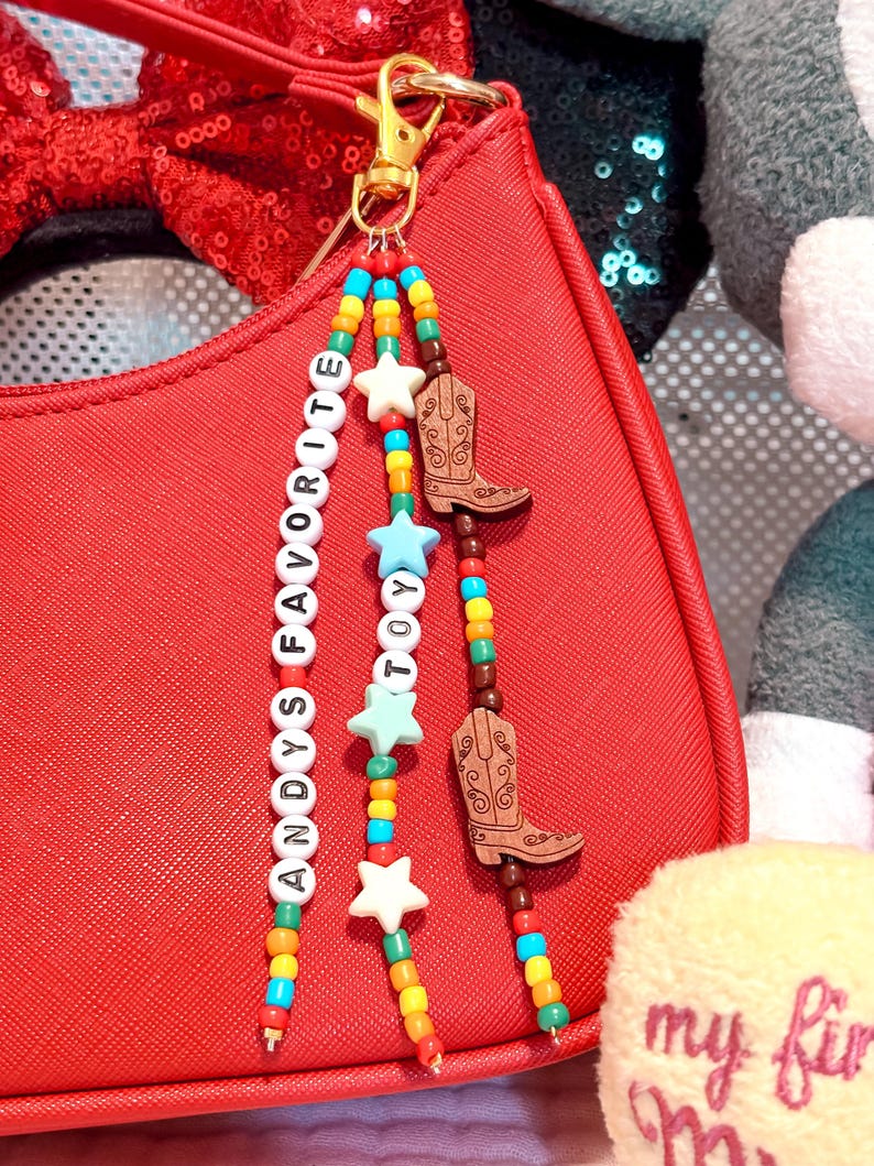 K&ouml;nnte beinhalten: Ein Taschenanh&auml;nger mit drei Perlenstr&auml;ngen an einem goldfarbenen Karabinerverschluss. Die Str&auml;nge bestehen aus bunten Kunststoffperlen, wei&szlig;en Buchstabenperlen mit der Aufschrift ANDYS FAVORITE TOY, hellblauen und wei&szlig;en sternf&ouml;rmigen Perlen sowie zwei braunen Holzanh&auml;ngern in Form von Cowboystiefeln.