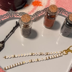May include: Two beaded keychains featuring white round beads with gold lettering and gold-tone spacers. One keychain reads ITS REALLY NOT SO SCARY and the other reads BOO TO YOU. Both include a gold-tone lobster clasp and are displayed on a heart-shaped tray.