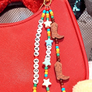 K&ouml;nnte beinhalten: Ein Taschenanh&auml;nger mit drei Perlenstr&auml;ngen an einem goldfarbenen Karabinerverschluss. Die Str&auml;nge bestehen aus bunten Rocailles, wei&szlig;en Buchstabenperlen mit der Aufschrift ANDYS FAVORITE, hellblauen Sternperlen und zwei braunen Cowboy-Stiefeln aus Holz.