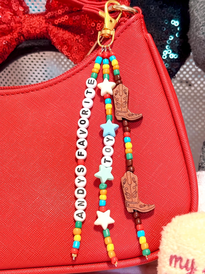 K&ouml;nnte beinhalten: Ein Taschenanh&auml;nger mit drei Perlenstr&auml;ngen an einem goldfarbenen Karabinerverschluss. Die Str&auml;nge bestehen aus bunten Rocailles, wei&szlig;en Buchstabenperlen mit der Aufschrift ANDYS FAVORITE, hellblauen Sternperlen und zwei braunen Cowboy-Stiefeln aus Holz.