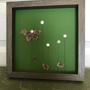 May include: Framed artwork featuring a green background with a floral design. The artwork includes clear and pink glass pieces, and white pearl-like accents. The frame is a weathered gray color.