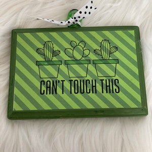 May include: A green wooden sign with a striped background and three potted cactus illustrations. The sign reads "Can't Touch This".