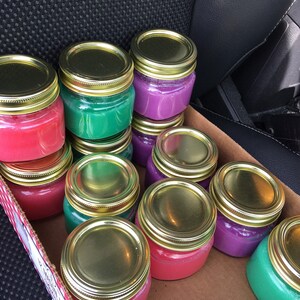 May include: A box filled with small glass jars with gold lids. The jars are filled with different colored wax, including red, purple, and green.