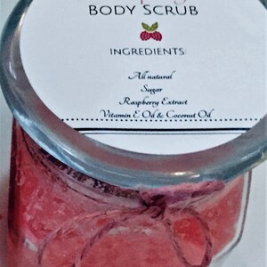 May include: A glass jar filled with a pink and white body scrub with a label that reads "Raspberry Body Scrub" and lists the ingredients as "All natural Sugar Raspberry Extract Vitamin E Oil & Coconut Oil".