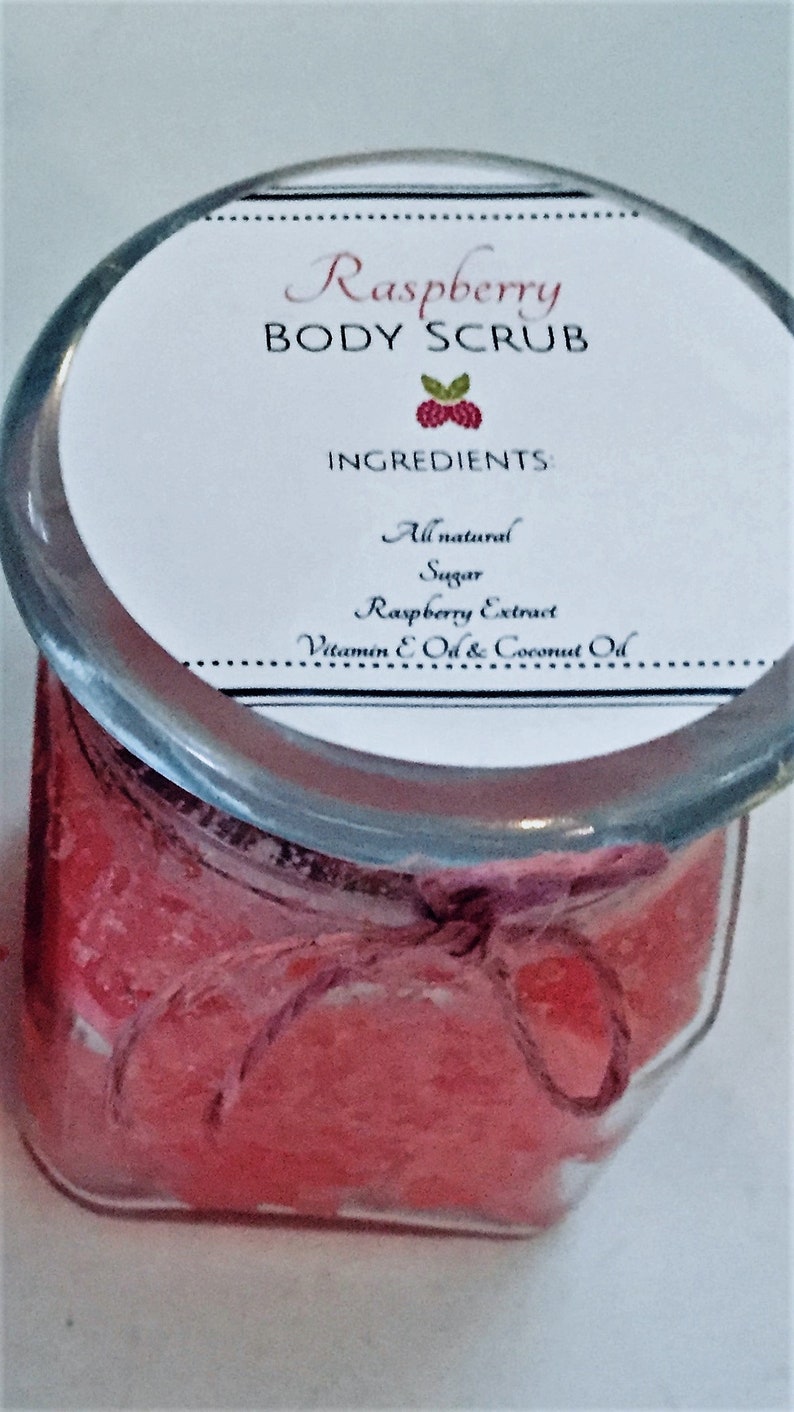 May include: A glass jar filled with a pink and white body scrub with a label that reads "Raspberry Body Scrub" and lists the ingredients as "All natural Sugar Raspberry Extract Vitamin E Oil & Coconut Oil".