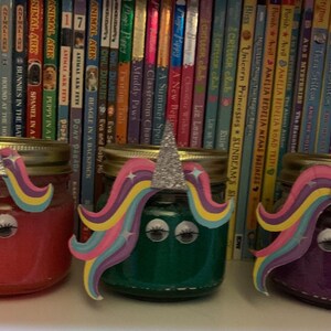 May include: Three glass jars decorated with unicorn faces. The jars are filled with pink, green, and purple glitter. The jars are sitting on a shelf in front of a bookshelf.